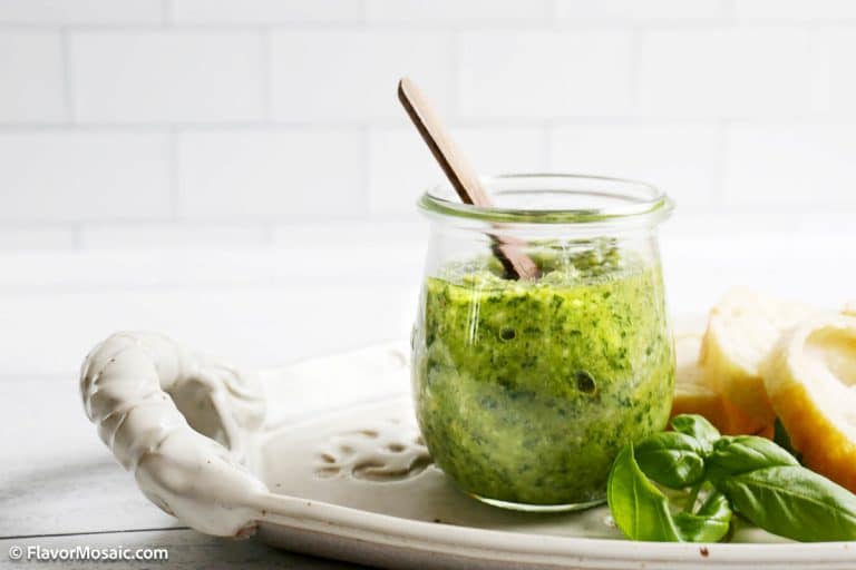 A glass jar of green pesto with a wooden spoon, placed on a white platter next to fresh basil leaves and lemon slices, perfect for elevating your favorite pesto recipe.
