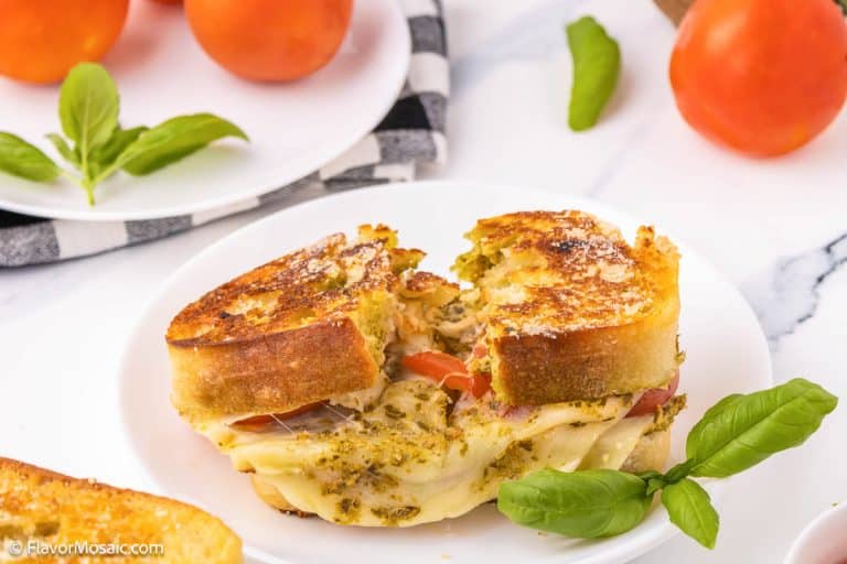 Tomato Pesto Grilled Cheese sandwich with melted cheese, pesto, and tomato slices on a white plate, garnished with fresh basil leaves. Tomatoes and basil sit in the background on a white surface.