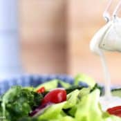 A close-up of creamy ranch dressing being poured from a glass pitcher onto a fresh salad with lettuce, cherry tomatoes, and cucumber slices.