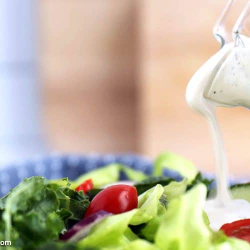 A close-up of creamy ranch dressing being poured from a glass pitcher onto a fresh salad with lettuce, cherry tomatoes, and cucumber slices.