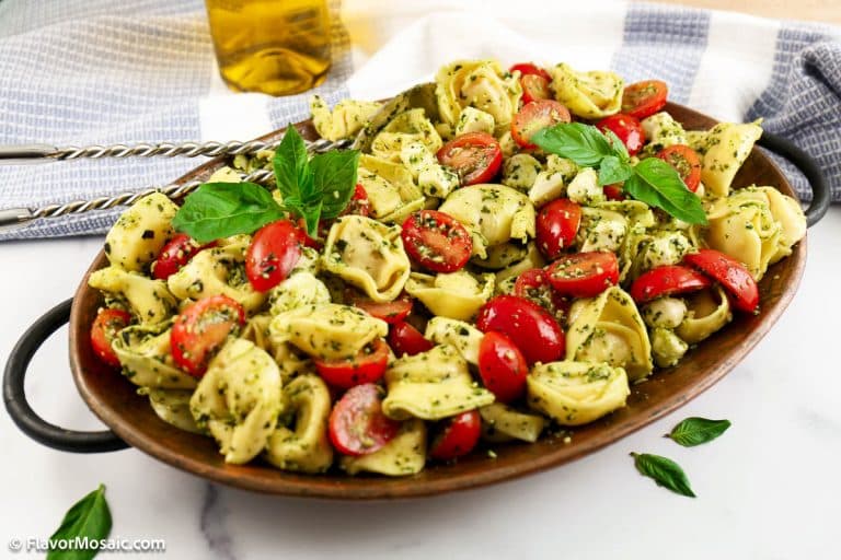 A platter of Pesto Tortellini Salad with cherry tomatoes, mozzarella, and pesto, garnished with fresh basil leaves. A pair of tongs and a bottle of olive oil are nearby on a striped towel.