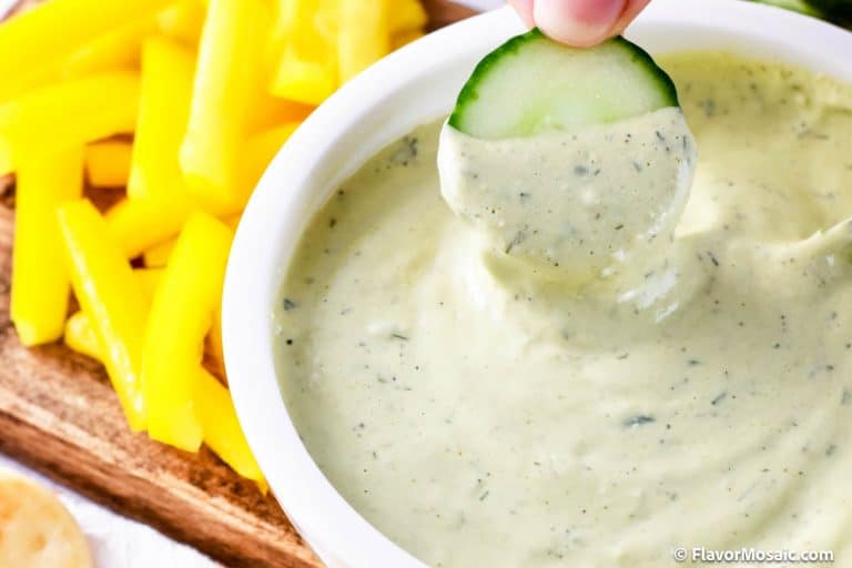 A cucumber slice is dipped into a creamy green Avocado Lime Ranch Dressing in a white bowl, with yellow bell pepper strips on the side.
