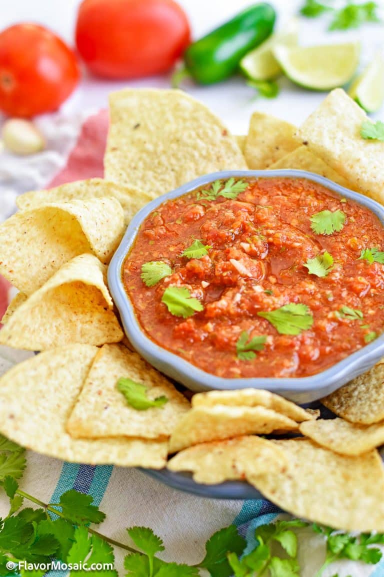 A bowl of red salsa topped with chopped fresh cilantro in a blue bowl surrounded by tortilla chips, with fresh cilantro, tomatoes, garlic cloves, jalapenos, and lime wedges surrounding the bowl.