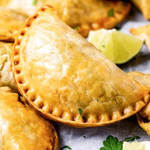 A single brisket empanada sitting on top of other beef brisket empanadas with a slice of lime and cilantro in the bottom right and a cut lime above the main empanada.