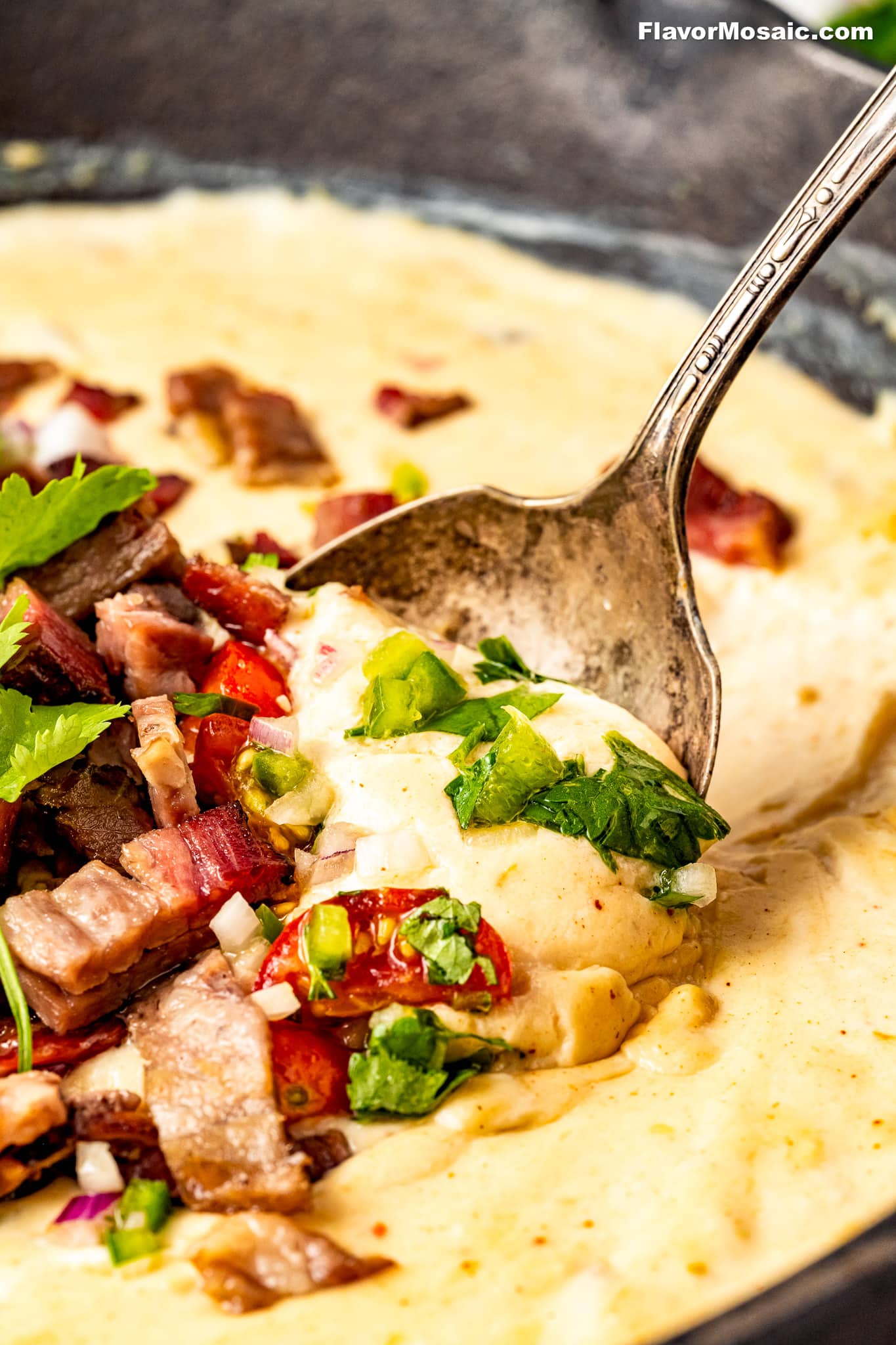 A spoon scoops creamy cheesy grits topped with chopped smoked meats, fresh cilantro, diced red peppers, and a drizzle of Brisket Queso from a bowl. The dish appears hearty and flavorful with colorful garnishes.