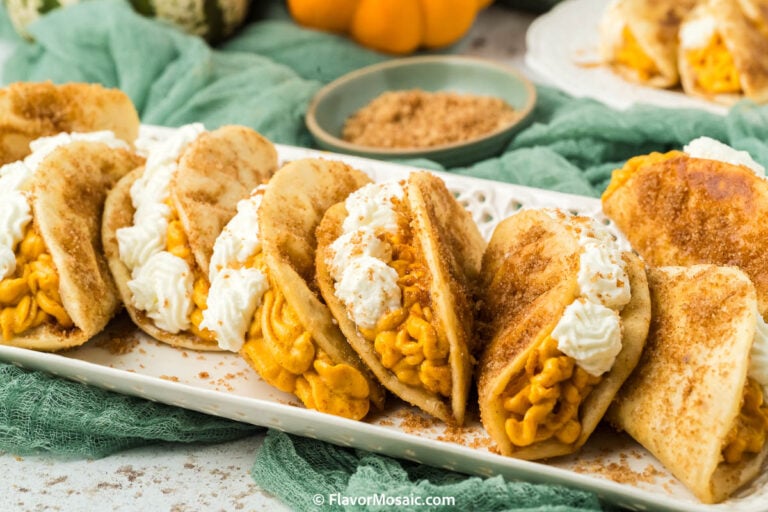 A plate of Pumpkin Pie Tacos filled with pumpkin cream and topped with whipped cream, sprinkled with cinnamon sugar. They are arranged in a row on a white platter with a green cloth and pumpkins in the background.