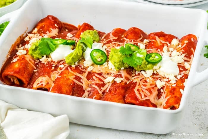 A white baking dish filled with chicken enchiladas rancheras, smothered in a homemade red enchilada sauce, topped with melted cheese, sour cream, guacamole, jalapeño slices, fresh cilantro, and crumbled cheese. A white napkin is nearby.