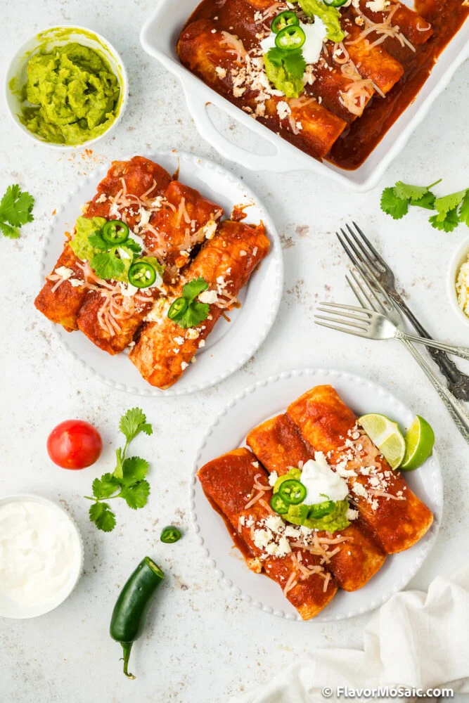 Overhead view of plates and a baking dish with cheesy enchiladas rancheras topped with sour cream, jalapeños, cilantro, and cheese. Sides of guacamole, lime wedges, a tomato, jalapeño, and forks are also visible.