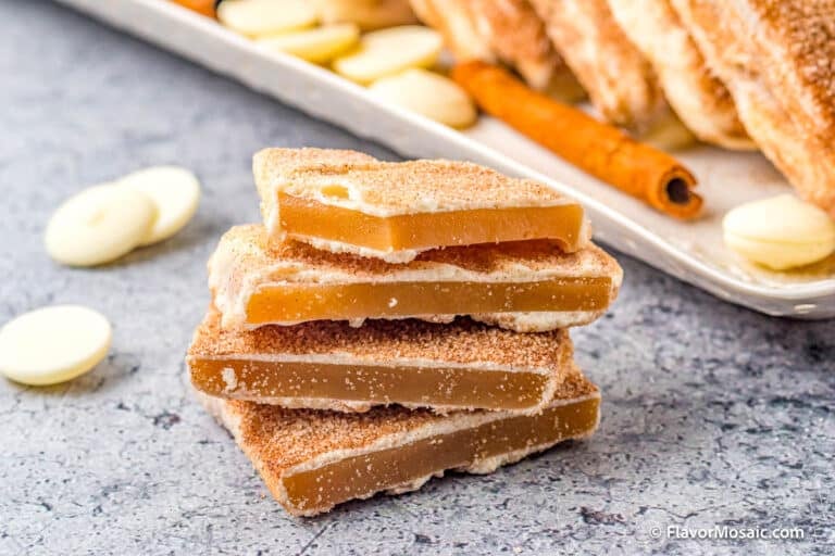 A stack of golden brown Churro Toffee pieces coated with cinnamon sugar, with white chocolate chips and cinnamon sticks in the background on a gray surface.