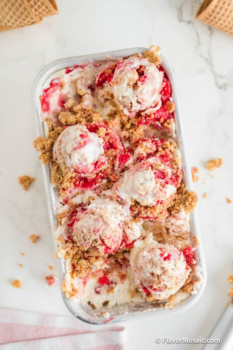 A container of Strawberry Cheesecake Ice Cream with several scoops on top, swirled with strawberry sauce and sprinkled with crumbly biscuit topping. Pieces of ice cream cone are visible in the background.