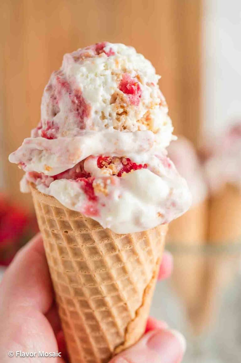 Someone holding a waffle cone with two scoops of Strawberry Cheesecake Ice Cream.