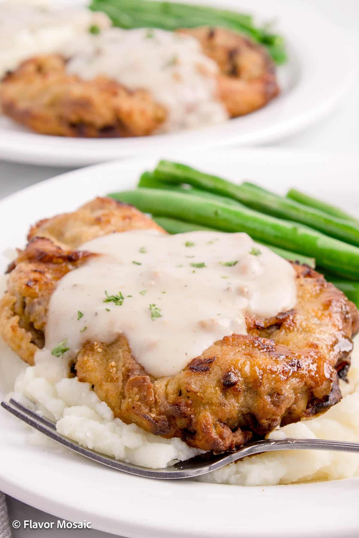 Air Fryer Chicken Fried Steak sitting on mashed potatoes on white plate with green beans next to it and another white plate in the background with the same dish.