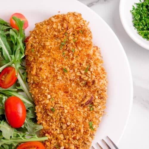 One piece of pecan crusted chicken breast on a white plate with a salad, and the end of a fork sitting on the plate.