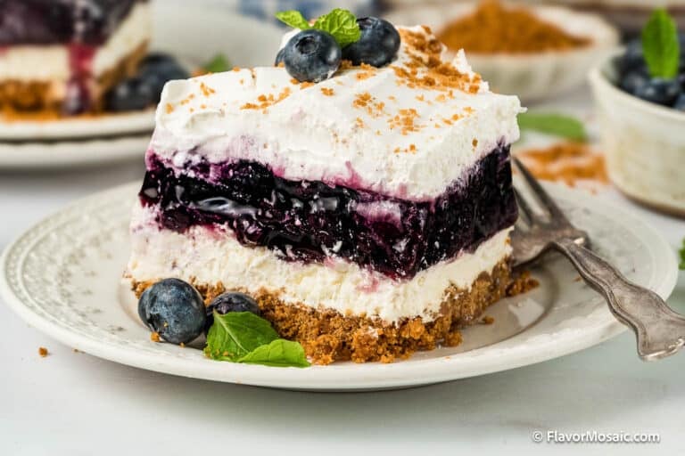 A slice of Blueberry Yum Yum cheesecake with a crumbly graham cracker crust, topped with whipped cream, fresh blueberries, and a mint leaf, served on a plate with a fork.
