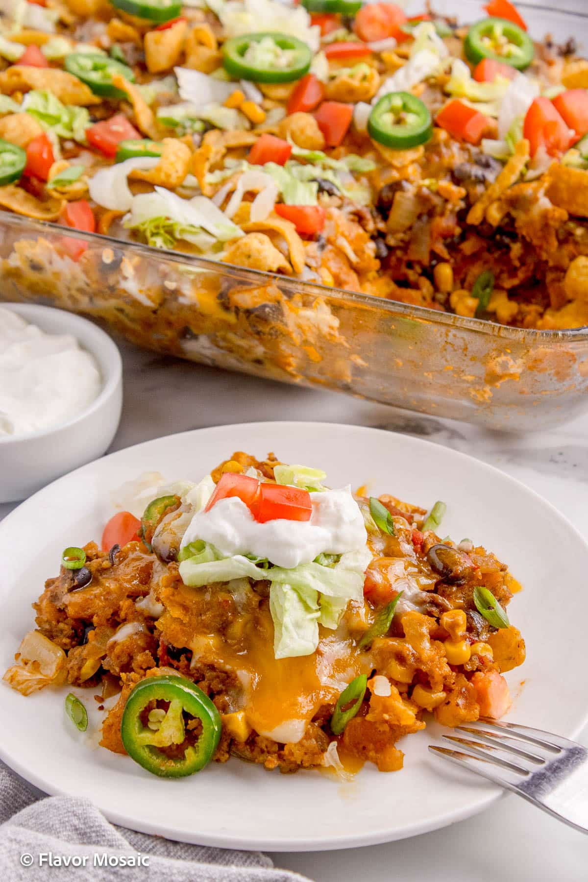 One serving of Frito Taco Pie Casserole, topped with sour cream, shredded lettuce, and chopped tomatoes, on a white plate with the rest of the casserole in the background.