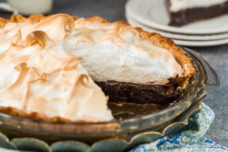 A classic Chocolate Meringue Pie in a glass dish, with golden, fluffy meringue topping and rich chocolate filling. A slice is missing, revealing its layers, while plates with another Chocolate Meringue Pie slice sit in the background.