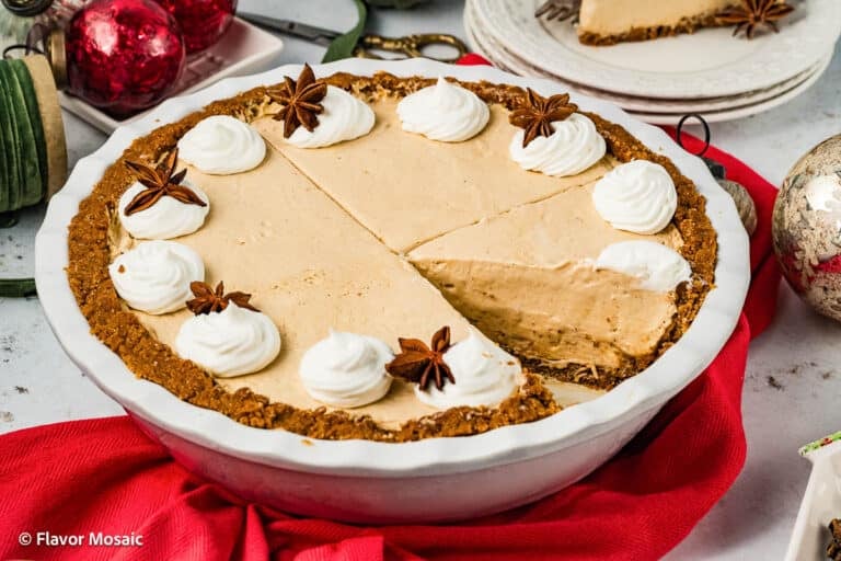 A creamy No Bake Eggnog Cream Pie with a graham cracker crust, topped with whipped cream and star anise, sits on a red cloth. One slice is missing, and holiday ornaments are nearby, adding festive cheer.