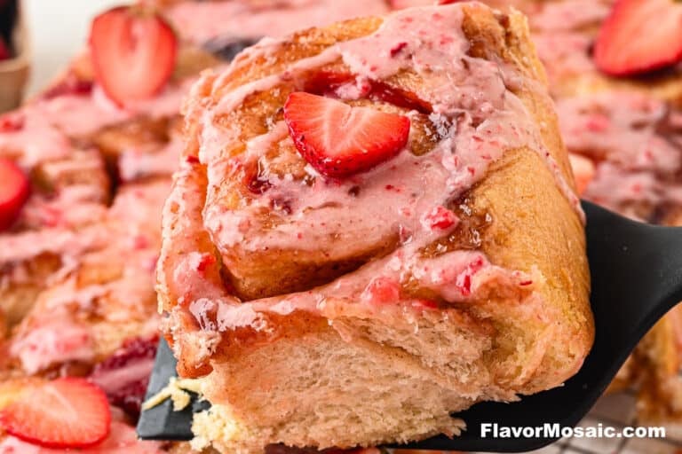 A close-up of a Strawberry Cinnamon Roll topped with pink strawberry glaze and a fresh strawberry slice, being lifted from a tray of Strawberry Cinnamon Rolls. The texture of the roll is soft and fluffy.