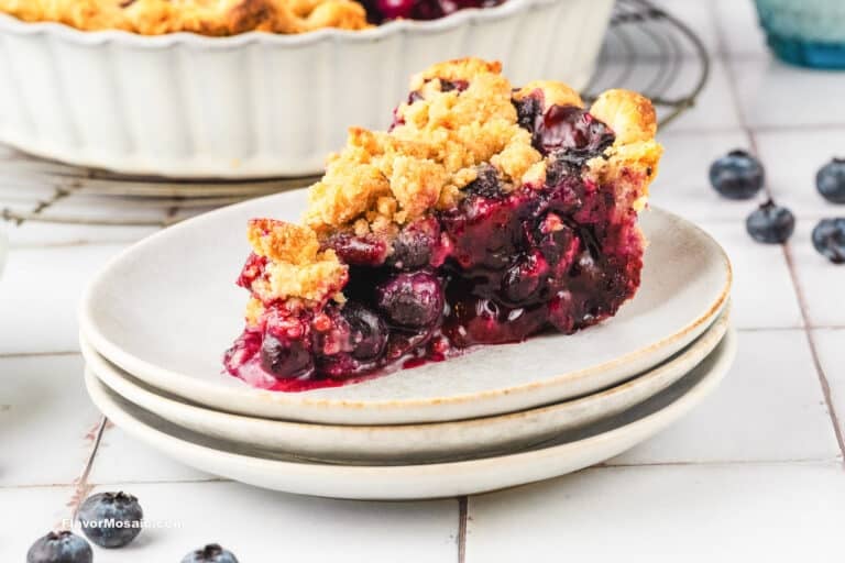 A slice of Blueberry Crumble Pie with a golden, crumbly topping sits on a stack of two white plates. Fresh blueberries and a pie dish are visible in the background.