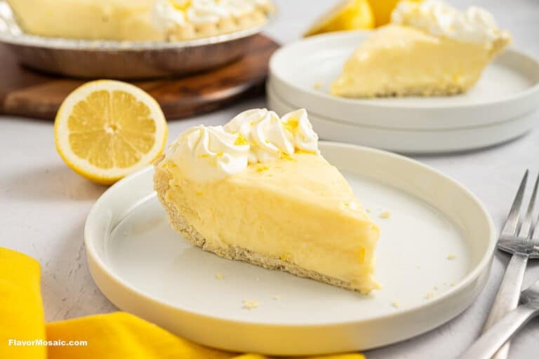 A slice of creamy Sour Cream Lemon Pie with whipped cream sits on a white plate, accompanied by a halved lemon and another pie slice in the background. A yellow napkin and two forks rest beside the plate.