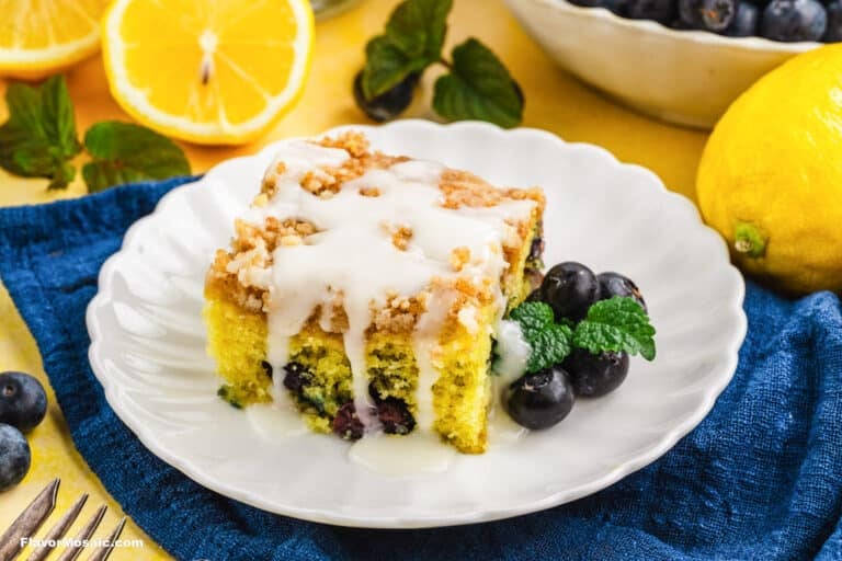 A slice of Lemon Blueberry Coffee Cake with crumb topping and white icing is served on a white plate, garnished with fresh blueberries and mint, with lemons and a bowl of blueberries in the background.