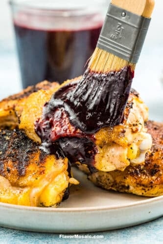 Brushing blueberry BBQ sauce on cooked chicken thighs with a jar of blueberry BBQ sauce in the background.
