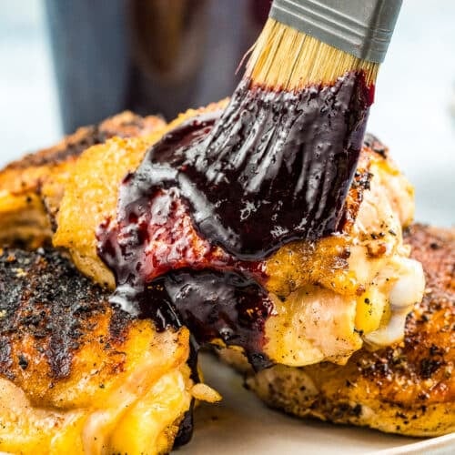 Brushing blueberry BBQ sauce on cooked chicken thighs with a jar of blueberry BBQ sauce in the background.