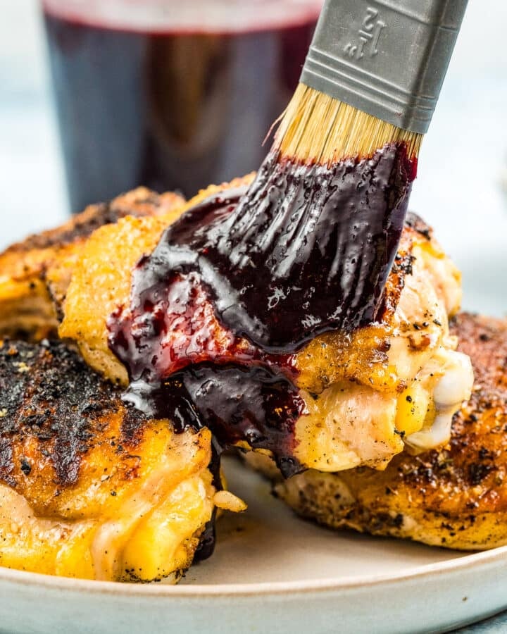 Brushing blueberry BBQ sauce on cooked chicken thighs with a jar of blueberry BBQ sauce in the background.