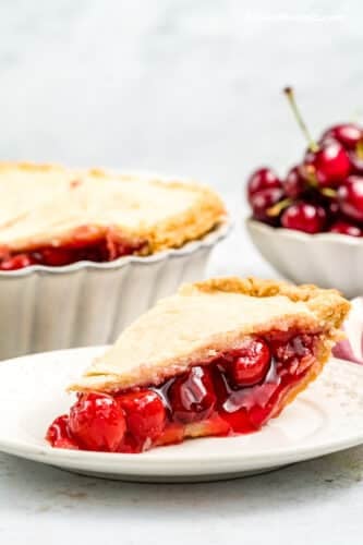 A side view of a slice of cherry almond pie on a white plate with the rest of the pie and a bowl of fresh cherries in the background.