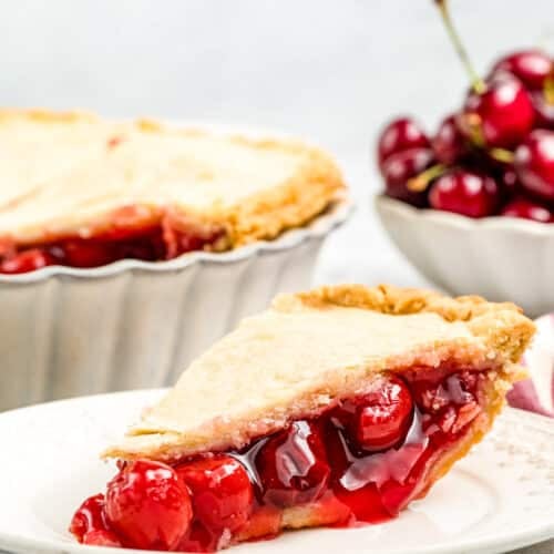A side view of a slice of cherry almond pie on a white plate with the rest of the pie and a bowl of fresh cherries in the background.