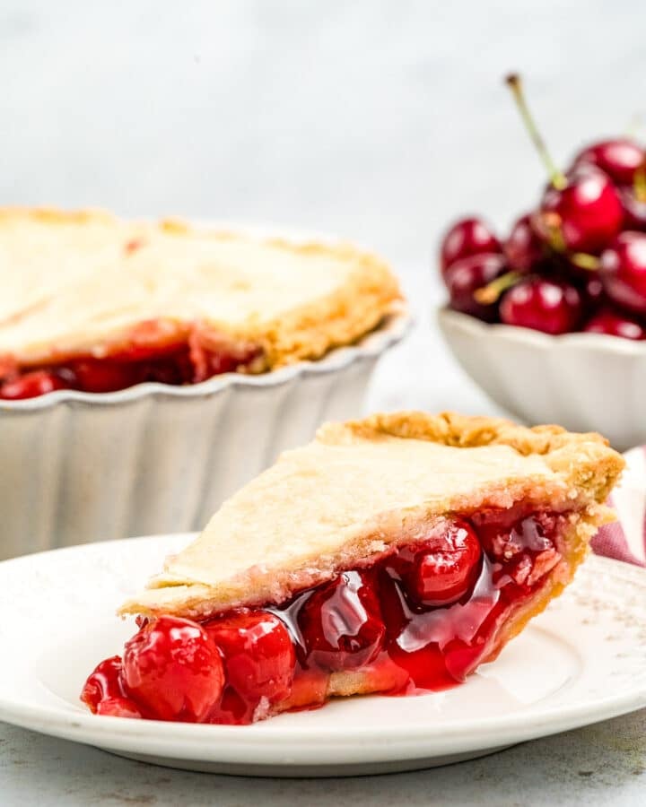 A side view of a slice of cherry almond pie on a white plate with the rest of the pie and a bowl of fresh cherries in the background.