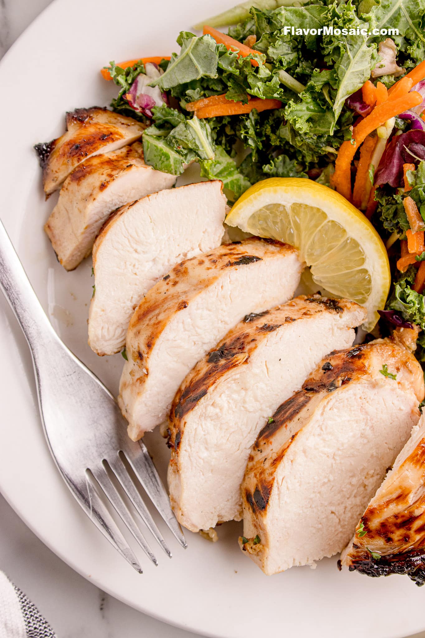 A juicy sliced chicken breast on a white plate with a green salad with shredded carrots, red cabbage, and a slice of lemon.