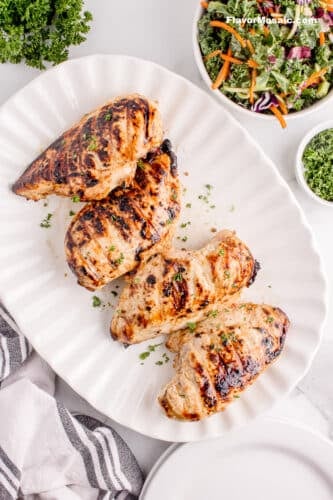 4 grilled chicken breasts on a white platter with a salad next to it.