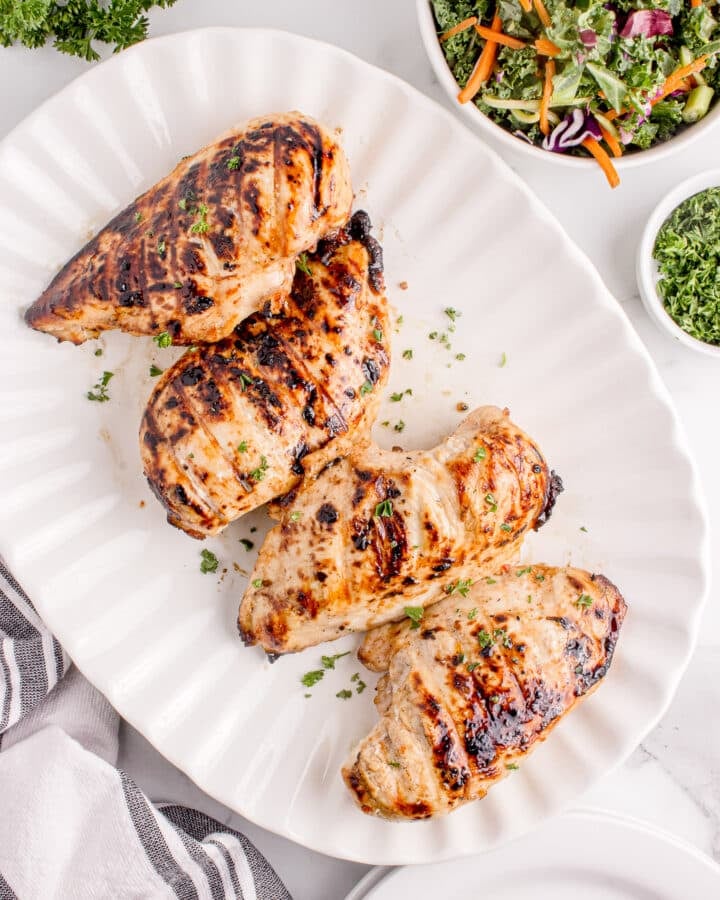 4 grilled chicken breasts on a white platter with a salad next to it.