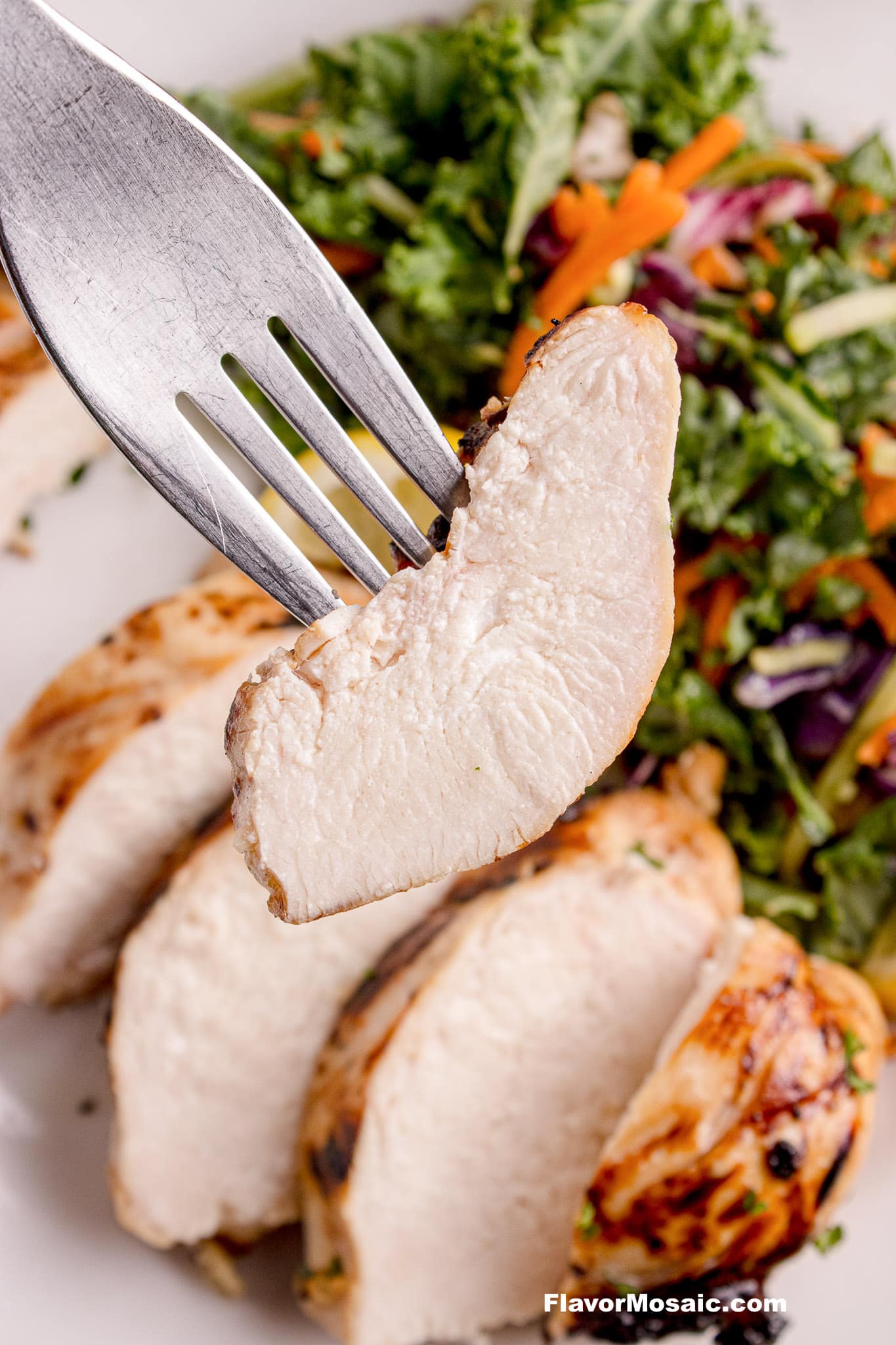 Fork holding a juicy, bite of moist grilled chicken breast. The rest of the sliced chicken breast is in the background with a green salad with shredded carrots and red cabbage.