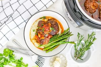 Overhead view of a serving of Instant Pot Braised Beef Short Ribs over mashed potatoes with asparagus with a partial view of the short ribs in the Instant Pot in the top right corner and fresh rosemary and parsley and minced garlic surrounding the bowl with the single serving.