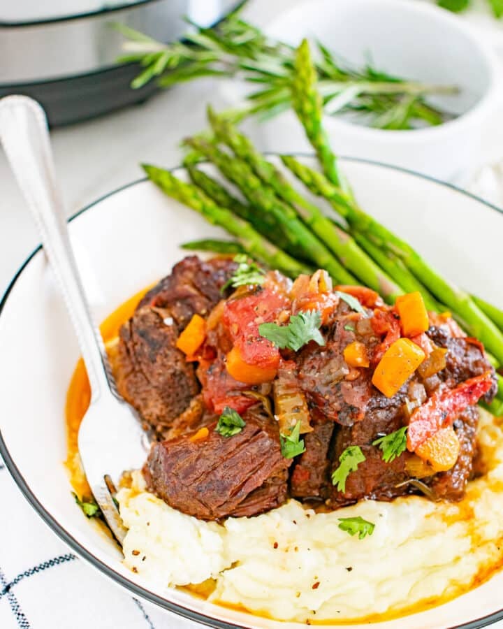 3/4 view of a serving of Instant Pot Braised Beef Short Ribs over mashed potatoes with asparagus covered in a au jus sauce.