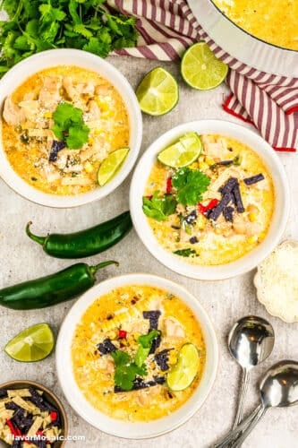 Three bowls of creamy Mexican Street Corn White Chicken Chili are garnished with lime wedges, cilantro, and tortilla strips. Jalapeños, fresh cilantro, lime halves, shredded cheese, and two spoons sit nearby atop a striped towel.