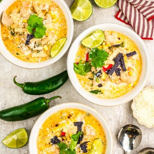 Three bowls of creamy Mexican Street Corn White Chicken Chili are garnished with lime wedges, cilantro, and tortilla strips. Jalapeños, fresh cilantro, lime halves, shredded cheese, and two spoons sit nearby atop a striped towel.