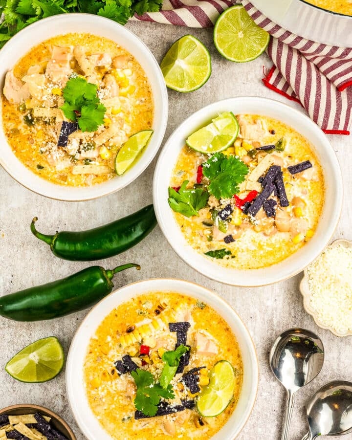 Three bowls of creamy Mexican Street Corn White Chicken Chili are garnished with lime wedges, cilantro, and tortilla strips. Jalapeños, fresh cilantro, lime halves, shredded cheese, and two spoons sit nearby atop a striped towel.