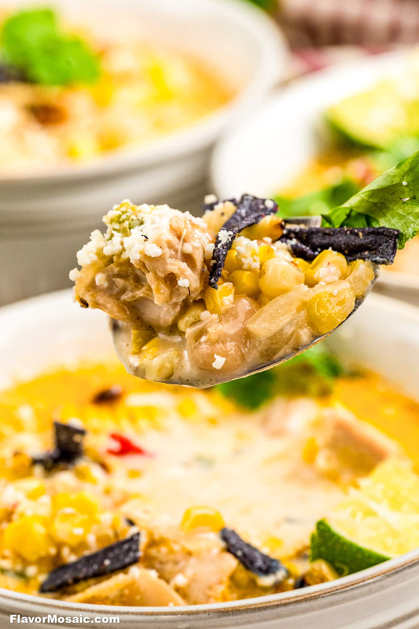 A close-up of a spoonful of creamy Mexican Street Corn White Chicken Chili soup, topped with crumbled cheese and tortilla strips, held above a bowl filled with the same hearty soup.