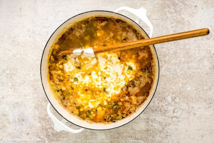 A pot of Mexican Street Corn White Chicken Chili with a wooden spoon, featuring vegetables and broth, is topped with a dollop of sour cream or yogurt and sits on a light-colored surface.