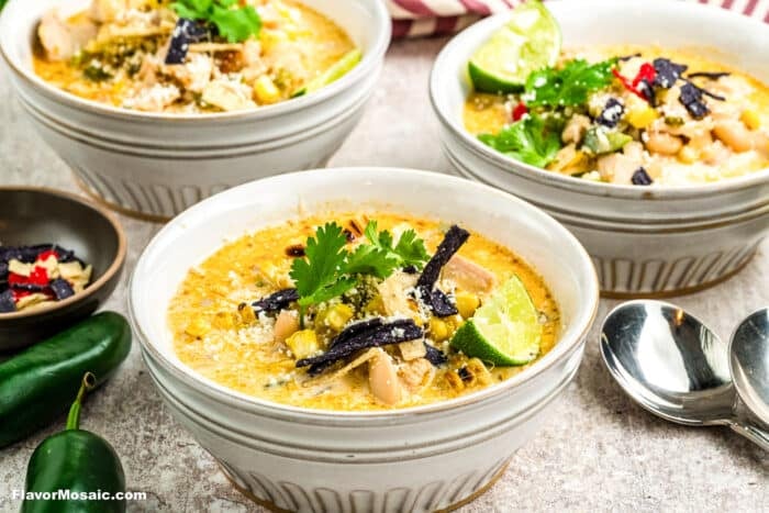 Three bowls of creamy Mexican Street Corn White Chicken Chili topped with cilantro, lime wedges, tortilla strips, corn, and cheese, with jalapeños and spoons nearby on a light surface.