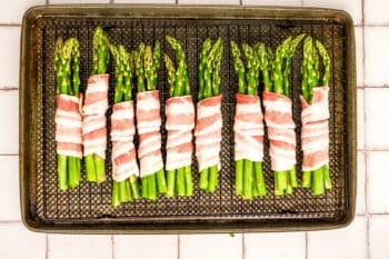 A baking tray with Bacon-Wrapped Asparagus bundles arranged in neat rows on a wire rack, ready to be cooked. The tray sits on a white tiled surface.