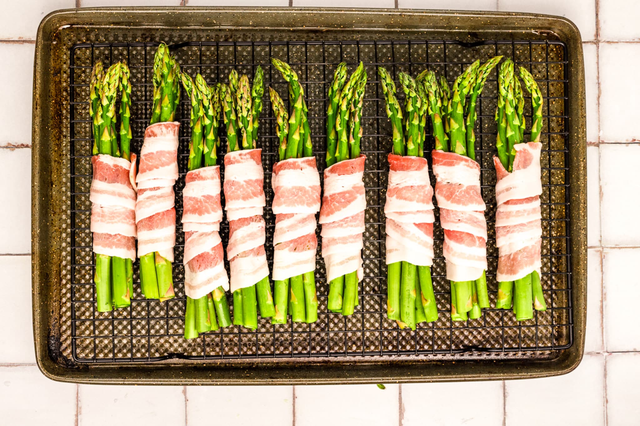 A baking tray with Bacon-Wrapped Asparagus bundles arranged in neat rows on a wire rack, ready to be cooked. The tray sits on a white tiled surface.