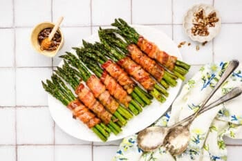 A platter of bacon-wrapped asparagus bundles sits on a white tiled surface, accompanied by a honey dipper in a small bowl, a dish of chopped pecans, two vintage spoons, and a floral napkin.