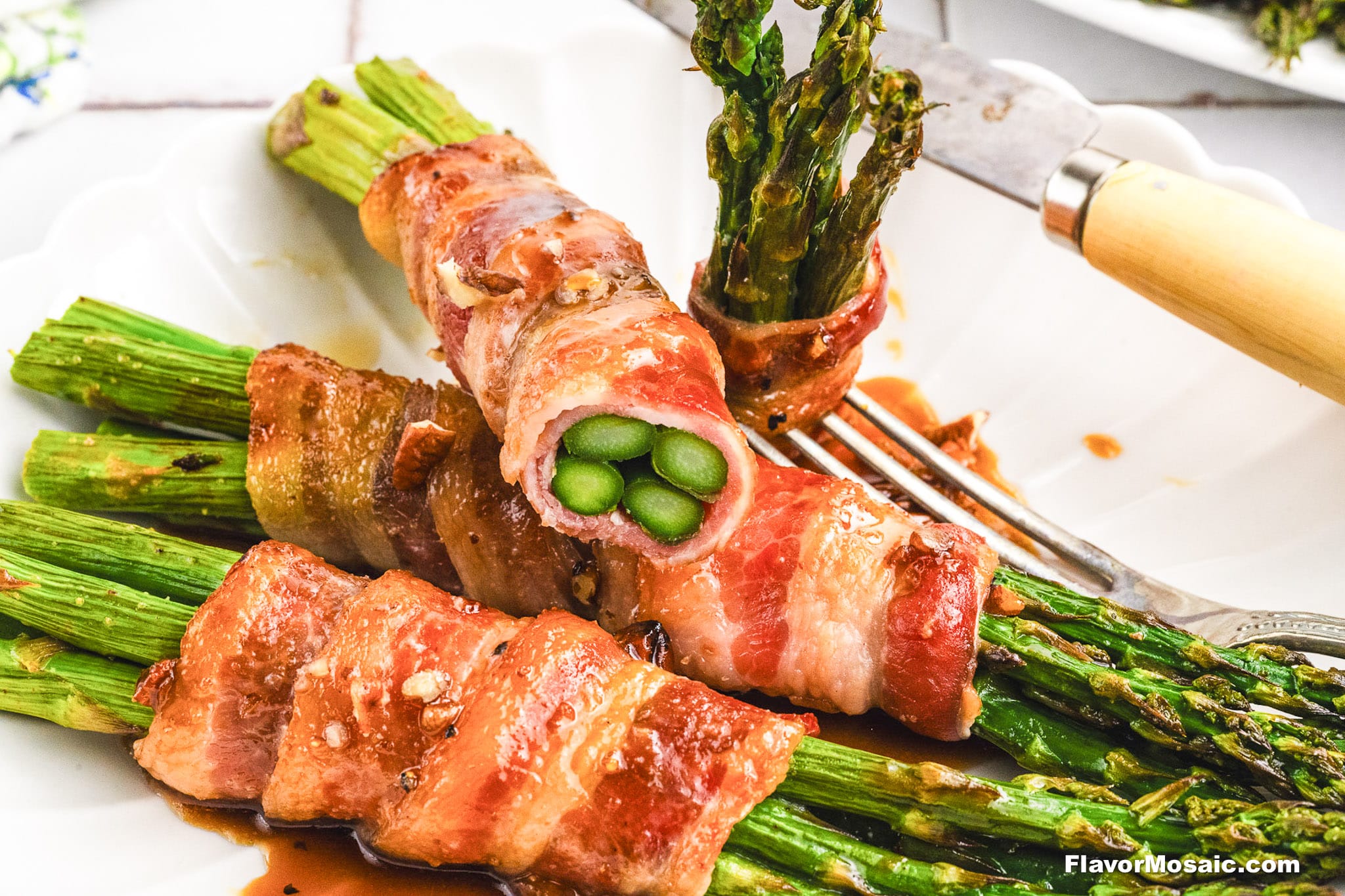 Three bacon-wrapped asparagus bundles are served on a white plate. A fork and knife rest nearby, with one Bacon-Wrapped Asparagus bundle cut open to reveal the vibrant green spears inside.