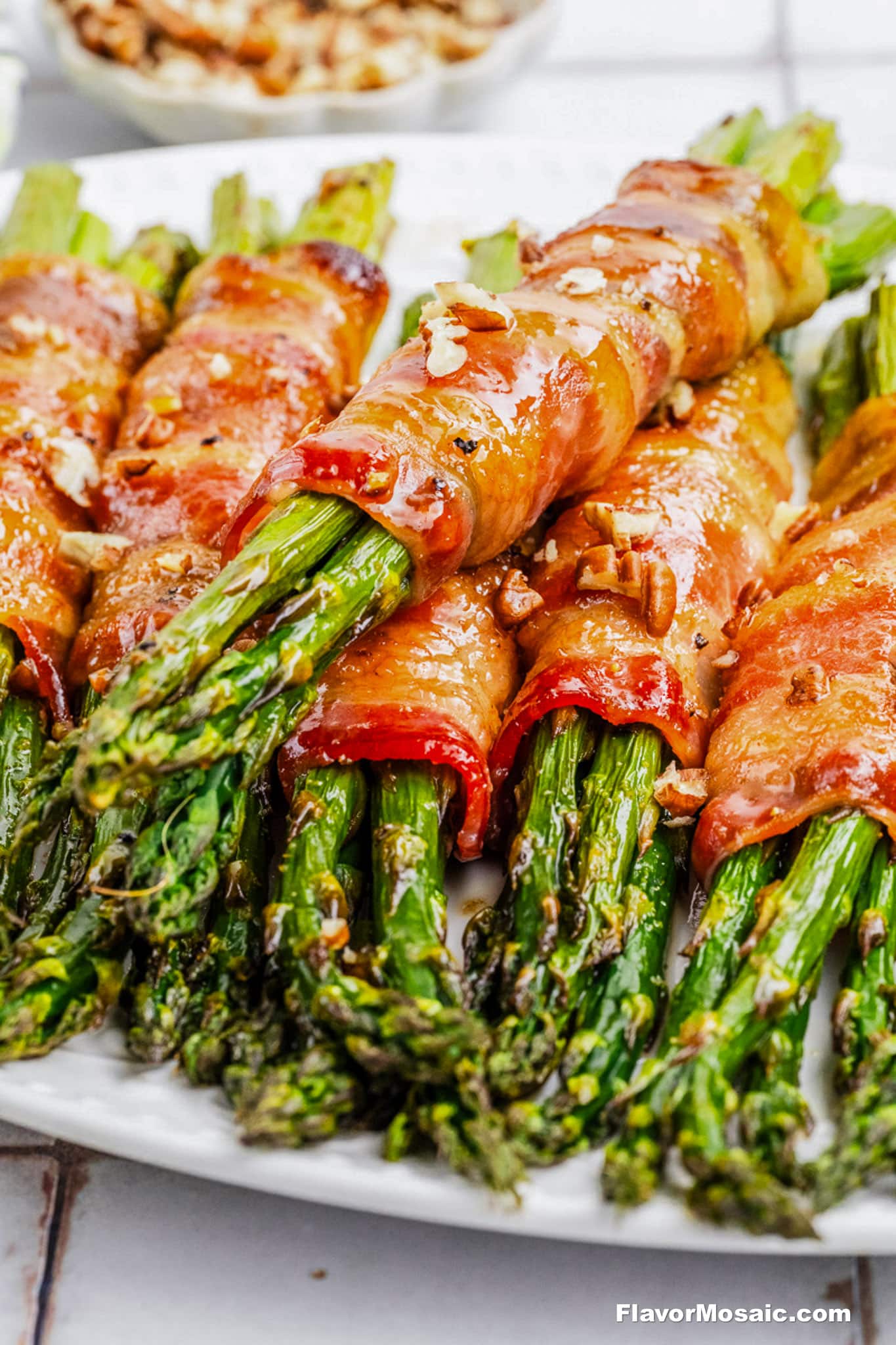 Close-up of bacon-wrapped asparagus bundles, garnished with chopped nuts and arranged on a white plate. The dish looks freshly cooked and ready to serve.