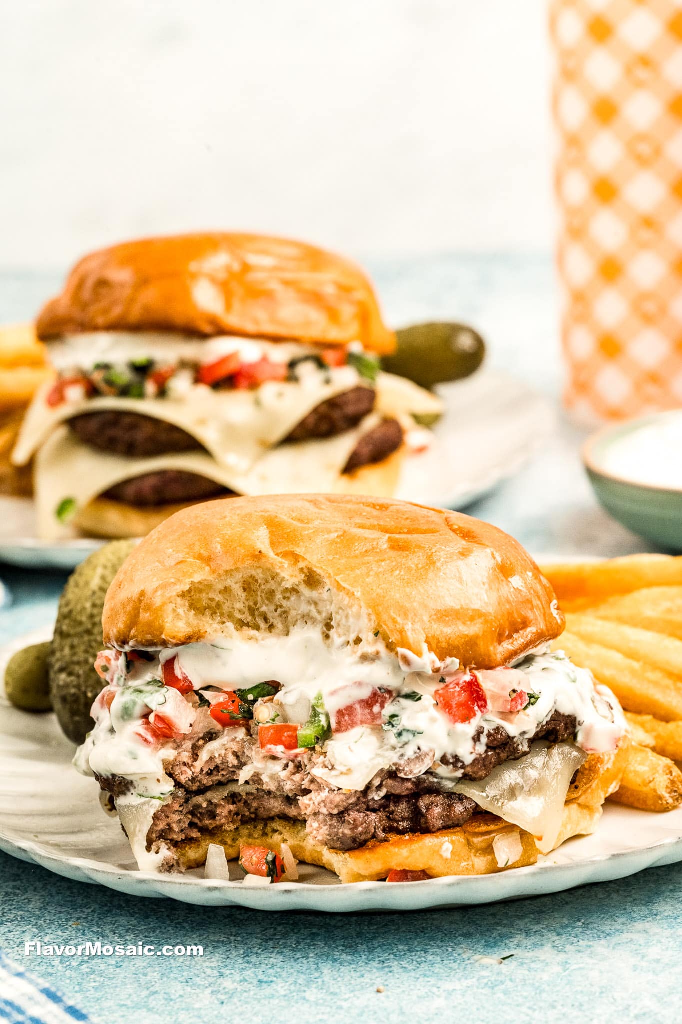 Two juicy double cheeseburgers topped with creamy sauce, diced tomatoes, and herbs sit on plates with crinkle-cut fries and pickles. The front burger has a bite taken out, revealing layers inspired by the Pico de Gallo Burger.