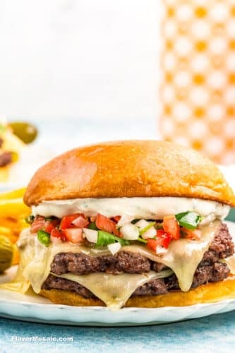 A cheeseburger with two beef patties, melted cheese, pico de gallo, and creamy sauce on a toasted bun, served on a white plate with crinkle-cut fries in the background.